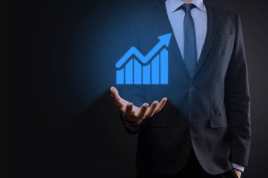 İş adamı elinde tablet tutuyor ve karanlık zemin üzerinde ok bulunan istatistik, grafik ve grafiklerin büyüyen sanal hologramını gösteriyor. Borsa. İş büyümesi, planlama ve strateji kavramı