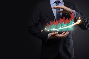 İş adamı elinde tablet tutuyor ve karanlık zemin üzerinde ok bulunan istatistik, grafik ve grafiklerin büyüyen sanal hologramını gösteriyor. Borsa. İş büyümesi, planlama ve strateji kavramı