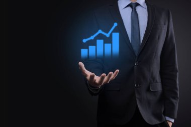 İş adamı elinde tablet tutuyor ve karanlık zemin üzerinde ok bulunan istatistik, grafik ve grafiklerin büyüyen sanal hologramını gösteriyor. Borsa. İş büyümesi, planlama ve strateji kavramı