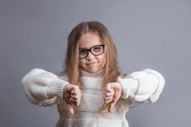 Süveter giyen, sarışın, kuşkucu, hoşlanmayan, onaylanmayan, gri arka planda iki eli olan genç ve çekici bir kızın portresi.