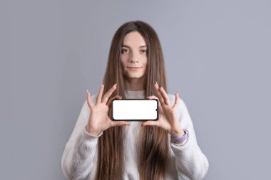 Süveter giyen, boş telefon ekranı gösteren mutlu, çekici esmer kadın. Kameraya bakıp gri arka planda gülümsemek..