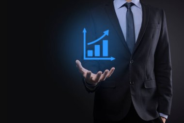 İş adamı elinde tablet tutuyor ve karanlık zemin üzerinde ok bulunan istatistik, grafik ve grafiklerin büyüyen sanal hologramını gösteriyor. Borsa. İş büyümesi, planlama ve strateji kavramı