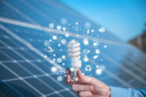 The man holds a light bulb, LED bulb on a background of solar panels with icons energy sources for renewable, sustainable development. Ecology concept