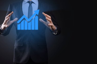 İş adamı elinde tablet tutuyor ve karanlık zemin üzerinde ok bulunan istatistik, grafik ve grafiklerin büyüyen sanal hologramını gösteriyor. Borsa. İş büyümesi, planlama ve strateji kavramı