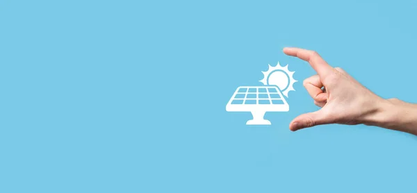 Hand on a blue background holds the icon symbol of solar panels. Renewable energy, solar panels station concept, green electricity.
