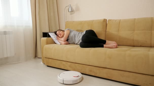 Brünette frau schlafen auf der couch roboter staubsauger reinigt den boden — Stockvideo