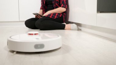 Robot elektrikli süpürge yerleri temizlerken telefonu olan tanınmayan bir kadın yerde oturuyor.