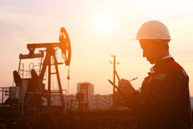 Beyaz kasklı bir adam gün batımında bir petrol pompasının arka planında bir kalemle kağıt dolduruyor.