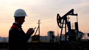 Beyaz miğferli, telefonlu bir adam sallanan bir petrol kuyusunun arka planında ve gün batımı gökyüzünde.