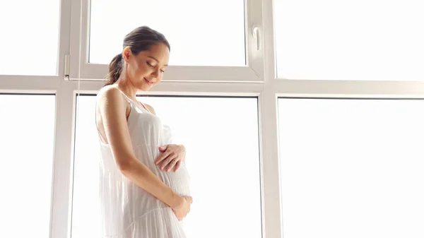 흰 옷을 입고 창문 가까이에서 배를 젓고 있는 임신부 — 스톡 사진