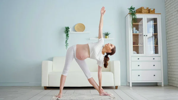 Mujer embarazada hace utthita trikonasana cerca de sofá en casa — Foto de Stock