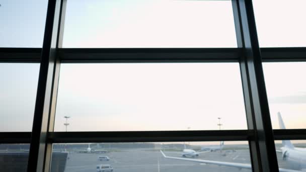 Mujer joven con una mochila se acerca a la ventana panorámica del aeropuerto con una vista de los aviones — Vídeo de stock