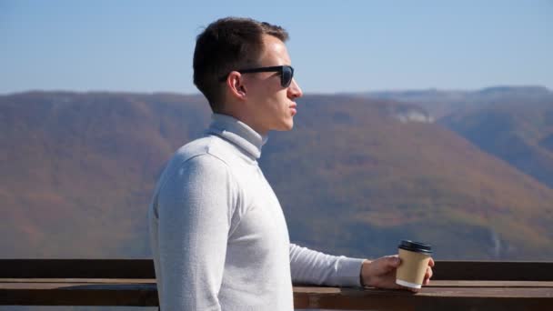 Junger Mann in grauem Rollkragen und Sonnenbrille trinkt an einem klaren Tag vor dem Hintergrund der Berge ein Getränk aus einem Plastikglas — Stockvideo