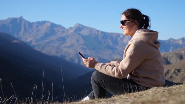 Mulher de óculos de sol e capuz senta-se no penhasco com telefone — Vídeo de Stock