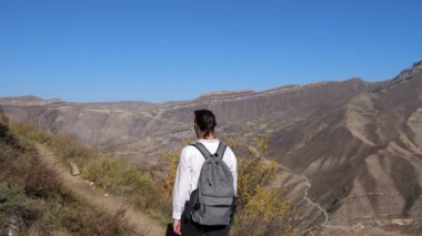 Sırt çantalı beyaz gömlekli genç bir kadın açık bir sonbahar gününde dağların arka planına doğru tırmanıyor.