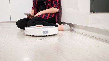 Robot elektrikli süpürge yerleri temizlerken telefonu olan tanınmayan bir kadın yerde oturuyor.