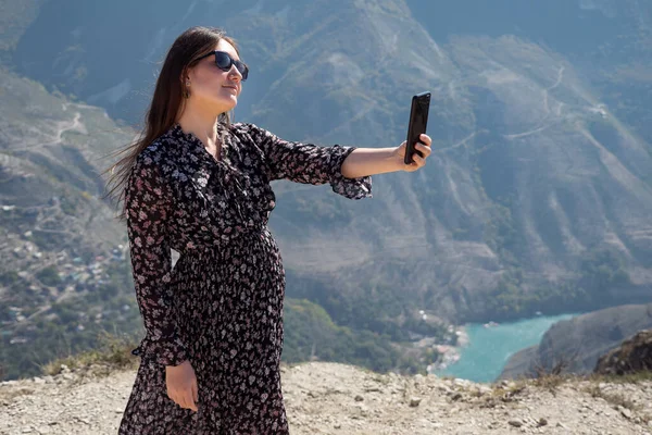Smiling lady in loose dress makes selfie against mountains