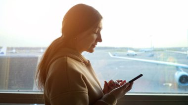 Uçak manzaralı pencerenin arka planında telefonu olan genç bir kadının yakın çekimi.