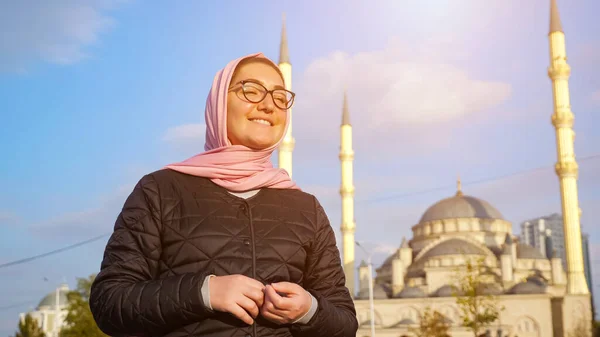 Νεαρή γυναίκα με ροζ μαντήλι και γυαλιά χαμογελώντας στο βάθος του τζαμιού — Φωτογραφία Αρχείου