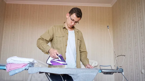 Junger Mann mit Brille und Hemd bügelt auf einem Bügelbrett — Stockfoto