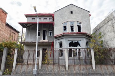 Yüksek çitlerin arkasında kırık camlı terk edilmiş iki katlı bir ev.