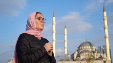 Pembe fularlı ve gözlüklü genç bir kadın ellerini caminin arka planında tutarken bir şeyler fısıldıyor.