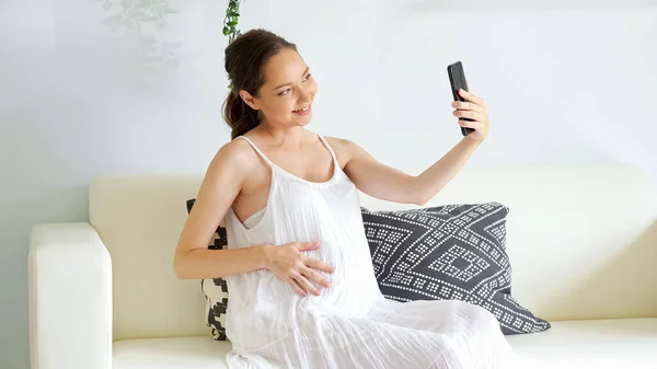 Pregnant woman has videocall with smartphone sitting on sofa — Stock Photo, Image
