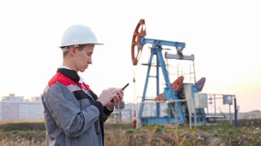 Beyaz kasklı bir adam, bir petrol kuyusunda sallanan arka planda telefonu var.