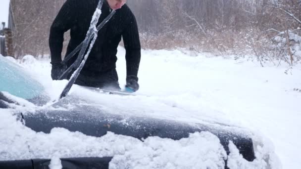 Man i svart jacka rengör en bil från snö under ett snöfall — Stockvideo