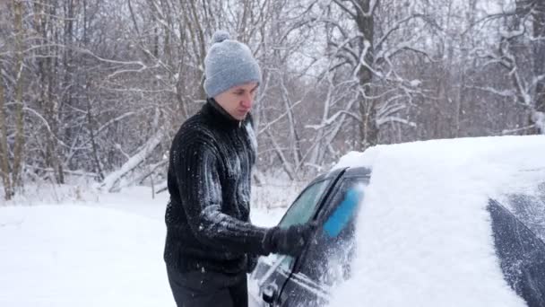黒いセーターと灰色の帽子の男がスローモーションで雪から車をきれいにする — ストック動画