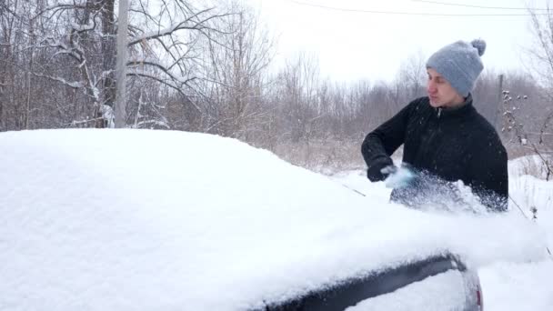 O homem em uma camisola preta limpa a janela traseira de um carro da neve durante uma queda de neve — Vídeo de Stock