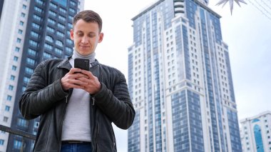 Deri ceketli bir adam telefonda uzun evlerin arka planında konuşuyor.