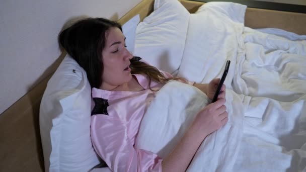 Lady encontra-se na cama segura smartphone lê e-book adormece — Vídeo de Stock