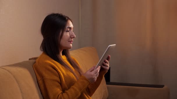 Koncentrerad brunett i gul tröja läser e-bok på soffan — Stockvideo