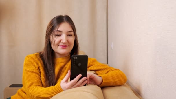 Giovane donna in maglione giallo siede sul divano marrone a leggere sms — Video Stock
