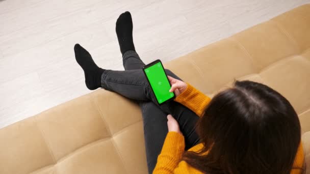 Брюнет у джинсах сидить на дивані з зеленим смартфоном. — стокове відео