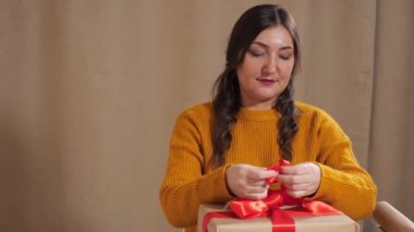 Kırmızı kurdeleyi yavaş çekimde bağlayan hediye kutusunu dekore eden genç bir kadın.