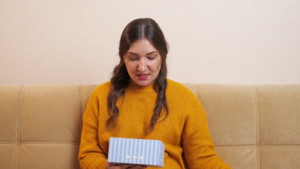 Schöne junge Frau sitzt auf dem Sofa und öffnet ein Geschenk und ist sehr enttäuscht — Stockvideo