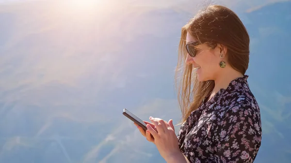 Fiatal nő telefonnal a háttérben a hegyek egy napsütéses napon, haj repked a szélben — Stock Fotó