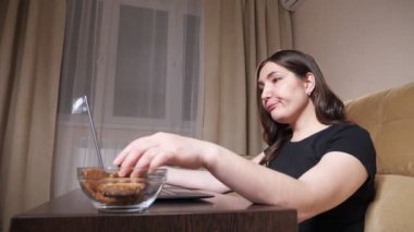 Siyah tişörtlü esmer, kurabiye yiyor ve dizüstü bilgisayarında yazı yazıyor.