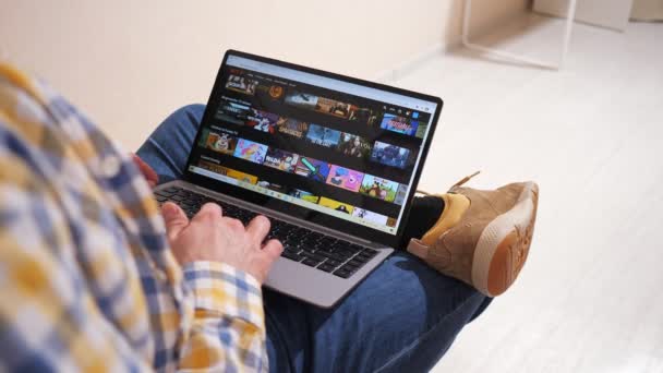 俄克拉荷马州卡赞- 2021年1月30日：一位身穿格子衬衫、头戴笔记本电脑的男子坐在客厅的沙发上，在Netflix上搜寻在线电影或电视剧 — 图库视频影像