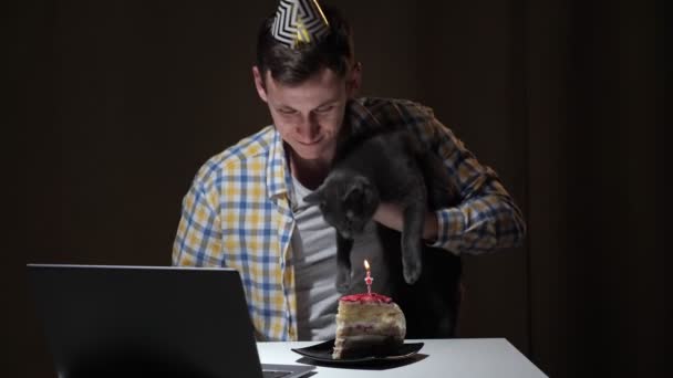 Chico en sombrero de cumpleaños de color se sienta con gato gris en la mesa con el ordenador portátil y saluda a los amigos en casa — Vídeos de Stock