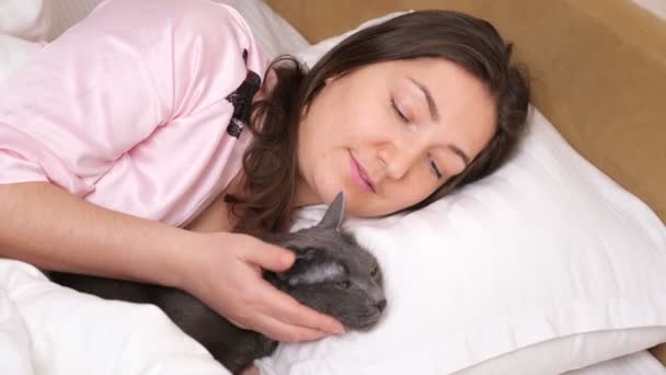Soluk pembe pijamalı rahat bir kadın. Yatakta uykulu bir kedi. — Stok video