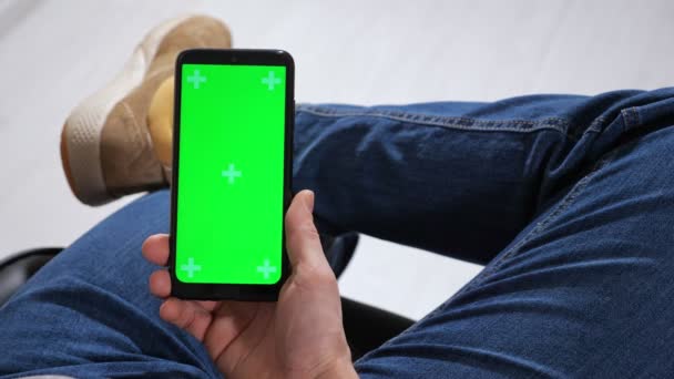 Onherkenbare man zit op een stoel met zijn benen gekruist over zijn knie en houdt een telefoon met een groen scherm in zijn handen — Stockvideo