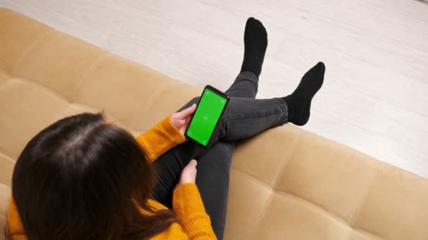 Brunette in jeans sits on sofa with green screen smartphone — Stock Video