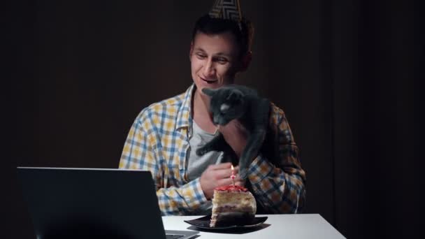 Kerel in gekleurde verjaardagshoed knuffelt grijze kat zittend aan tafel — Stockvideo