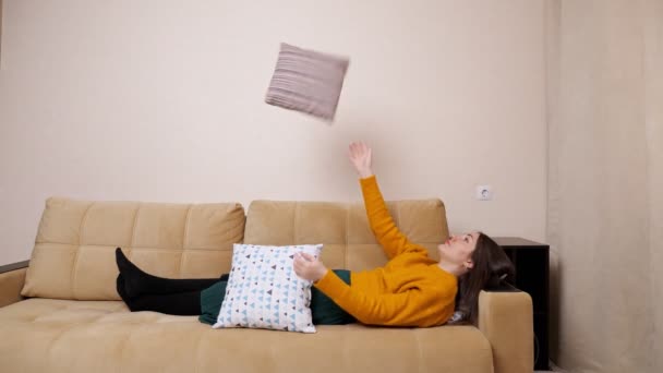 Набридла молода жінка в светрі лежить на дивані, кидаючи подушки — стокове відео
