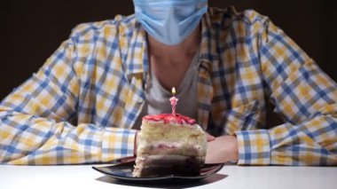 Tek kullanımlık mavi maskeli tanınmayan bir adam evde tek başına oturan bir tabak dolusu pastaya ateş üflüyor..