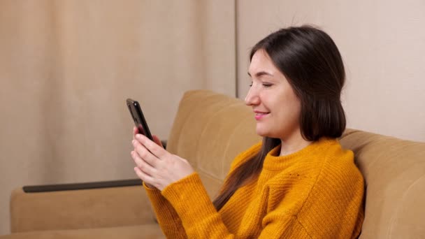 Brunette in pullover sits on sofa and types on smartphone — Stock Video