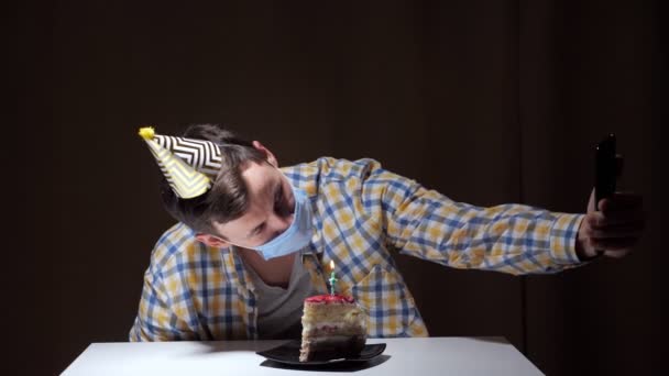 일회용 마스크 파티 모자를 쓴 남자가 케이크와 함께 하얀 테이블에 앉아 있는 검은 스마트폰에서 사진을 찍고 있습니다. — 비디오